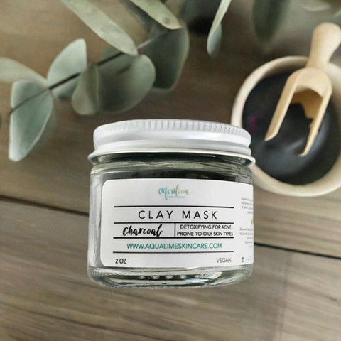 Aqualime Charcoal Clay Mask in a 2 oz glass bottle, accompanied by a bowl of activated charcoal and a fresh green leaf, highlighting the natural ingredients for detoxifying skin