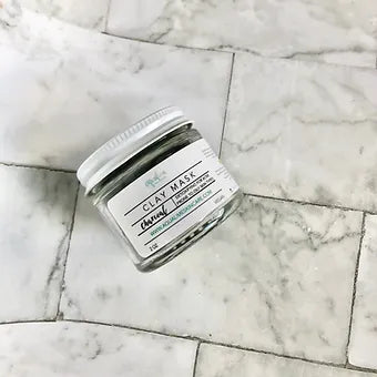 Aqualime Charcoal Clay Mask in a 2 oz glass bottle placed on white, soft flooring, highlighting its sleek design and natural detoxifying ingredients for clear skin
