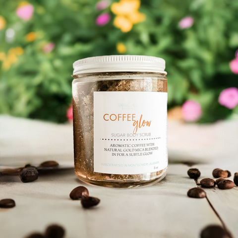 Aqualime Coffee Glow Shimmering Sugar Body Scrub in a glass bottle placed on a white surface, surrounded by coffee seeds, with a green leaf and yellow flower in the background, emphasizing its natural ingredients for glowing skin