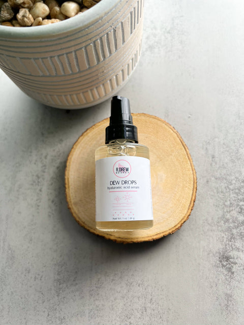 R. Drew Dew Drops Hyaluronic Acid Serum bottle placed on a cutting wooden plate with a pot on a floral background, enhancing hydration and soothing