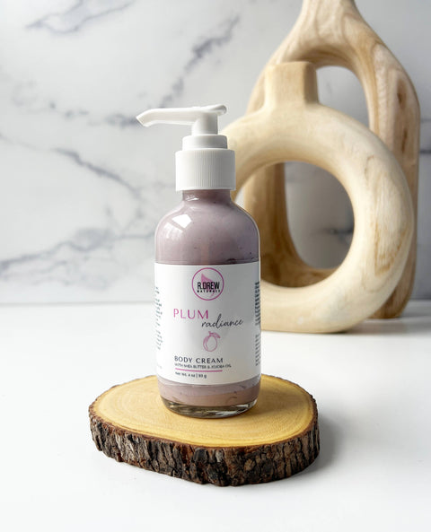 R. Drew Plum Radiance Shea Butter Body Cream bottle with round design placed on a wooden plate with fresh background, offering hydration and a sugar plum fragrance