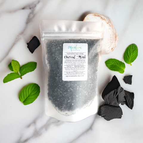 A sleek black pouch of Aqualime Charcoal & Mint Detox Soaking Salt, surrounded by activated charcoal pieces, fresh peppermint leaves, and Epsom salts on a white marble background, highlighting its natural ingredients and luxurious feel.