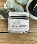 Aqualime Charcoal Clay Mask in a 2 oz glass bottle placed on a wooden surface, with a bowl of activated charcoal and a fresh green leaf, showcasing natural ingredients for detoxifying skin