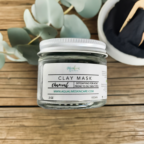 Aqualime Charcoal Clay Mask in a 2 oz glass bottle placed on a wooden surface, with a bowl of activated charcoal and a fresh green leaf, showcasing natural ingredients for detoxifying skin