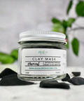 Aqualime Charcoal Clay Mask in a 2 oz glass bottle, placed on a white background surface with activated charcoal and a fresh green leaf, highlighting its natural ingredients for skin detoxification