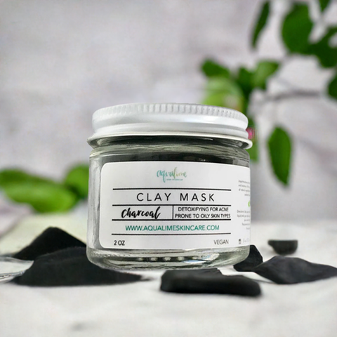 Aqualime Charcoal Clay Mask in a 2 oz glass bottle, placed on a white background surface with activated charcoal and a fresh green leaf, highlighting its natural ingredients for skin detoxification