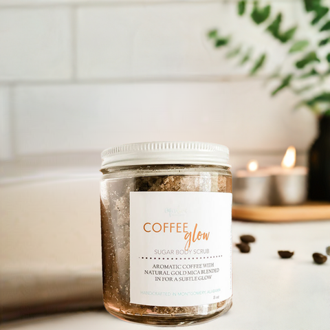 Aqualime Coffee Glow Shimmering Sugar Body Scrub in a glass bottle, surrounded by coffee seeds and a fresh green leaf, highlighting its aromatic blend of coffee, cinnamon, and gold mica for a glowing skin