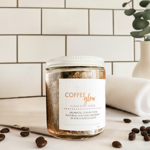 Aqualime Coffee Glow Shimmering Sugar Body Scrub in a glass bottle placed on a white cloth background, with fresh coffee seeds and leaves surrounding it, highlighting its natural ingredients for glowing skin