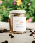 Aqualime Coffee Glow Shimmering Sugar Body Scrub in a glass bottle placed on a white surface, surrounded by coffee seeds, with a green leaf and yellow flower in the background, emphasizing its natural ingredients for glowing skin