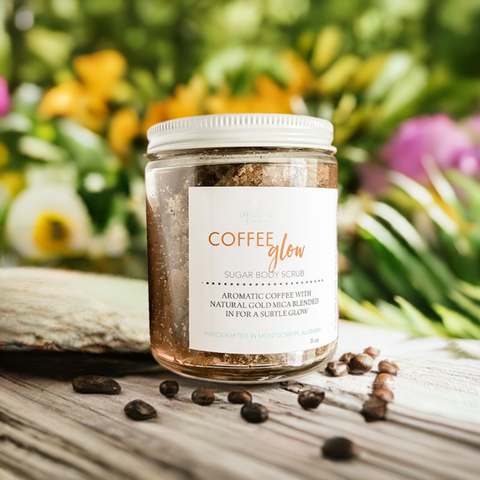 Aqualime Coffee Glow Shimmering Sugar Body Scrub in a glass bottle placed on a wooden surface, surrounded by coffee seeds, with a background of green leaves, yellow and pink flowers, highlighting its natural ingredients for glowing skin