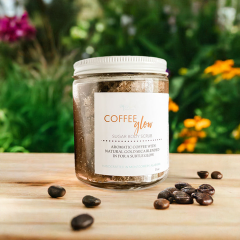 Aqualime Coffee Glow Shimmering Sugar Body Scrub in a glass bottle placed on a wooden surface, surrounded by coffee seeds, with green leaves and yellow and red flowers in the background, highlighting its natural and aromatic ingredients for glowing skin.