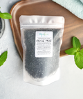 A sleek black pouch of Aqualime Charcoal & Mint Detox Soaking Salt placed on a wooden tray, surrounded by fresh peppermint leaves, highlighting its natural ingredients like activated charcoal, peppermint, and Epsom salts for a luxurious detox experience