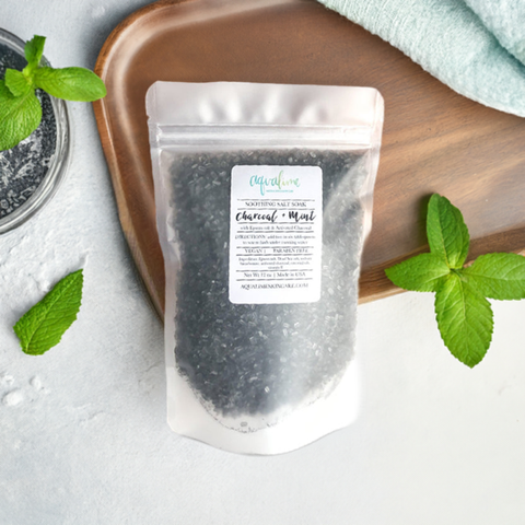 A sleek black pouch of Aqualime Charcoal & Mint Detox Soaking Salt placed on a wooden tray, surrounded by fresh peppermint leaves, highlighting its natural ingredients like activated charcoal, peppermint, and Epsom salts for a luxurious detox experience