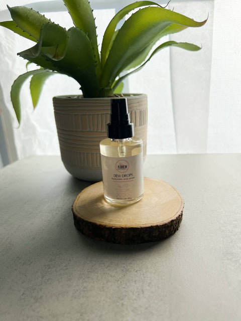 R. Drew Dew Drops Hyaluronic Acid Serum bottle placed on a wooden plate with an aloe vera pot on a white floor, promoting hydration and soothing