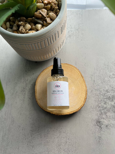 R. Drew Dew Drops Hyaluronic Acid Serum bottle placed on a cutting wooden plate with a green plant pot, offering hydration and soothing benefits